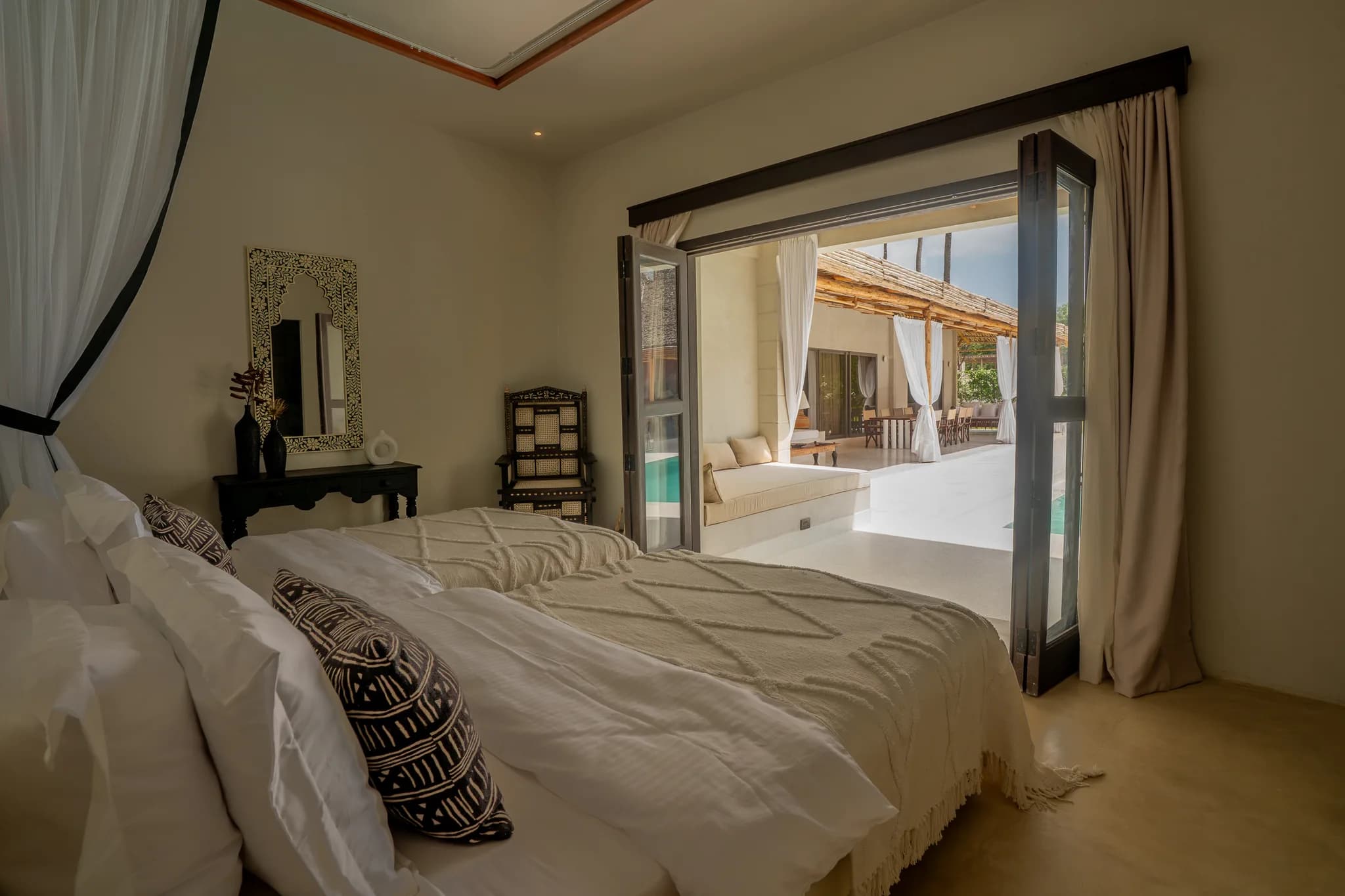 Bedroom with large bed draped in beige linens and open doors to poolside